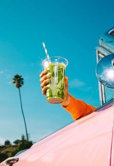 a person holding up a green drink in front of a pink car with palm trees