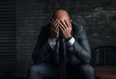 a man in a suit sitting on a chair covering his face with both hands while covering his eyes