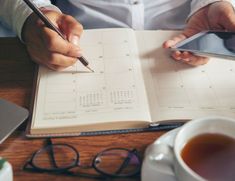 a person holding a pen and writing on a notebook with a cup of coffee next to it