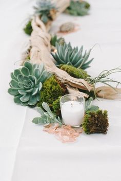 an arrangement of succulents and greenery on a white table cloth with a candle