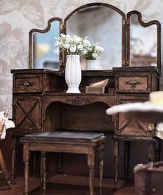 an antique dresser with mirror, stool and flowers in vase on table next to chair