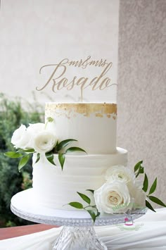 a white wedding cake with gold lettering and flowers on the top is sitting on a table