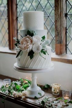 a three tiered cake with flowers on top is sitting on a table next to candles and greenery