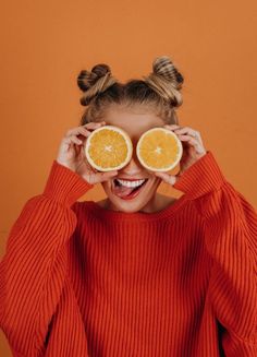 a woman holding two halves of oranges up to her eyes with both hands and smiling