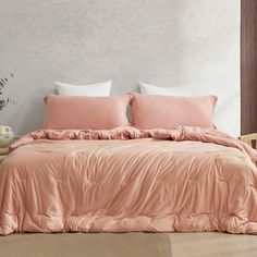 a bed with pink comforter and pillows in a white room next to a potted plant