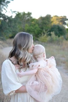 a woman holding a baby in her arms while standing on a dirt road with trees in the background