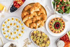 a white table topped with lots of plates and bowls filled with food next to croissants