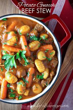 a red pot filled with stew and carrots on top of a wooden table next to a spoon