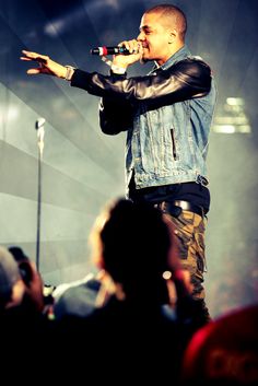 a man standing on top of a stage holding a microphone in his hand and wearing a jean jacket