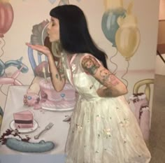 a woman in a white dress blowing on a piece of cake with balloons behind her