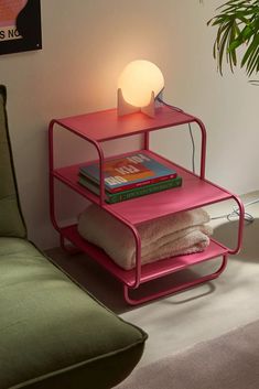 a pink shelf next to a green couch with books on it and a lamp in the corner