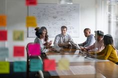 a group of people sitting around a conference table with sticky notes on the wall behind them