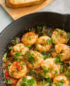 a skillet filled with shrimp and vegetables on top of a wooden cutting board next to bread