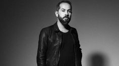 a man with a beard standing in front of a gray wall wearing a leather jacket