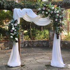 an outdoor wedding setup with white draping and flowers on the arch, surrounded by greenery