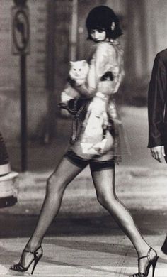 a black and white photo of a woman walking down the street with a cat in her hand