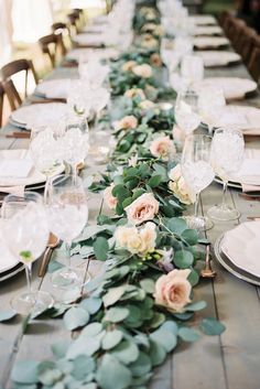 a long table is set with white plates and silverware, greenery and flowers