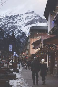 two people are walking down the street in front of some buildings with snow covered mountains behind them