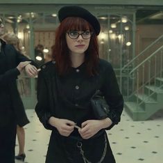 a woman with red hair and glasses is standing in a room holding a handbag