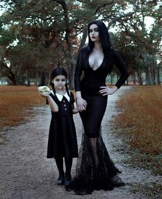 two women in black dresses standing next to each other on a dirt road with trees and grass behind them