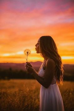 a woman in a white dress holding a dandelion with the sun setting behind her