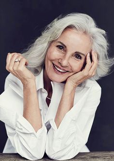 an older woman sitting at a table with her hands on her chin and smiling for the camera