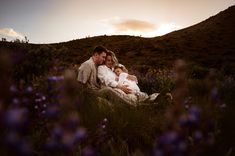 a man and woman are sitting in the grass with their baby on their lap as the sun sets behind them