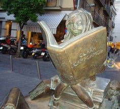 a statue of a girl reading a book in the middle of a busy city street