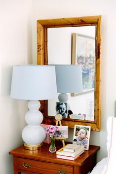 a nightstand with a lamp and pictures on it in front of a mirror above the bed