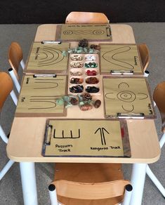 a wooden table topped with lots of different pieces of art on top of white chairs