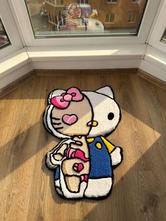 a hello kitty rug on the floor in front of a window with a cat holding a teddy bear