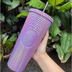 a hand holding a purple tumbler with a lion on it's side and green plants in the background