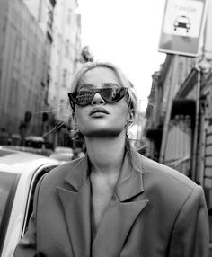 a woman wearing sunglasses standing in front of a car on a city street next to tall buildings