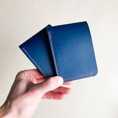 a hand holding two blue leather wallets on top of a white surface with red stitching