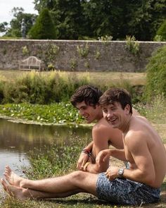 two young men sitting next to each other on the ground near water and grass, smiling