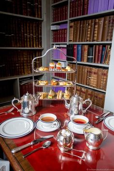 a red table topped with white plates and cups filled with tea next to bookshelves