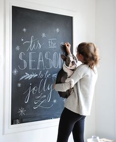 a woman writing on a blackboard with a dog in her lap and the words tis season for jollyy written on it