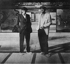 two men standing next to each other in front of a building