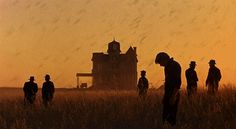 several people standing in a field watching the sun go down on an old farm house