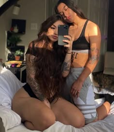 two beautiful women sitting on top of a bed next to each other holding a cell phone