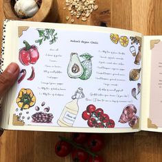 an open recipe book on a table with tomatoes, peppers, and other food items