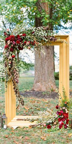 an arch decorated with greenery and red flowers sits in front of a large tree