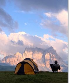 a man sitting in front of a tent on top of a lush green field next to a tall mountain