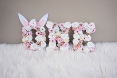 the word easter spelled with flowers and bunny ears on a fluffy white surface in front of a wall