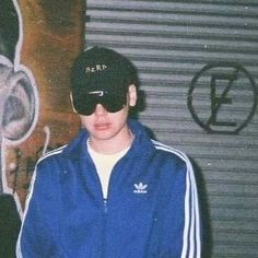 a man wearing a hat and sunglasses standing in front of a wall with graffiti on it