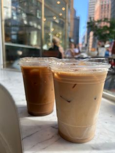 two iced coffees sitting on top of a table next to each other in plastic cups