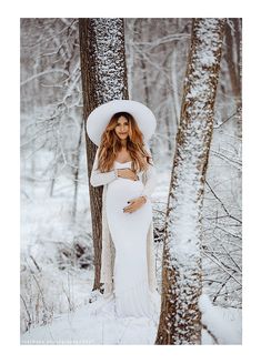 a pregnant woman wearing a white dress and hat in the snow with trees behind her