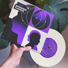 a person holding up two vinyl records in front of a plant with purple and black designs on it