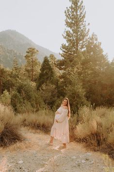 a pregnant woman standing on a dirt road in the middle of some brush and trees