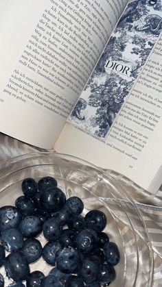 blueberries are in a glass bowl next to an open book on a table top